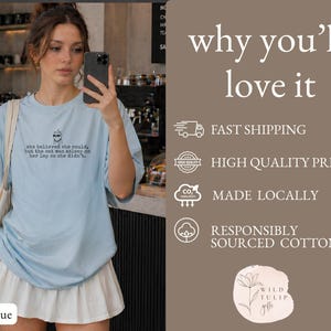 May include: A light blue t-shirt featuring a small graphic of a cat wearing sunglasses and the text: she believed she could, but the cat was asleep on her lap so she didn't. The shirt is paired with a white pleated skirt and a beige tote bag.