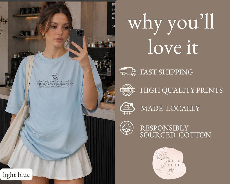 May include: A light blue t-shirt featuring a small graphic of a cat wearing sunglasses and the text: she believed she could, but the cat was asleep on her lap so she didn't. The shirt is paired with a white pleated skirt and a beige tote bag.