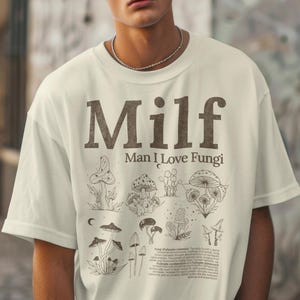 May include: Cream-colored t-shirt with the text "Milf Man I Love Fungi" in brown. The shirt features various mushroom illustrations in brown. A detailed description of fungi is printed below the illustrations.