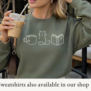 May include: An olive green sweatshirt with a white graphic of a coffee cup, cat, and book. The sweatshirt is being worn by a person holding an iced coffee. The text at the bottom says "Sweatshirts also available in our shop."