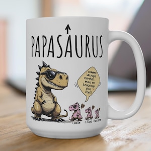 Puede incluir: Taza de cer&aacute;mica blanca con el texto "PAPASAURUS" y una flecha hacia arriba. La taza presenta un dinosaurio de dibujos animados con gafas de sol y tres dinosaurios m&aacute;s peque&ntilde;os. El texto en la taza dice "comme un papa normal mais en beaucoup plus cool!" y los nombres "Lucie, Chlo&eacute;, Roxane."