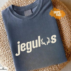 May include: Dark heather gray t-shirt with the word "Jeguls" in white, featuring a deer antler graphic. The shirt is displayed on a woven surface. The text "Ships Fast" is in an orange banner.