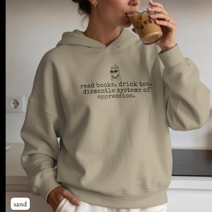 May include: A sand-colored hooded sweatshirt featuring a black graphic of a cat wearing sunglasses and a party hat, accompanied by the text: read books. drink tea. dismantle systems of oppression. The hoodie has long sleeves and ribbed cuffs.