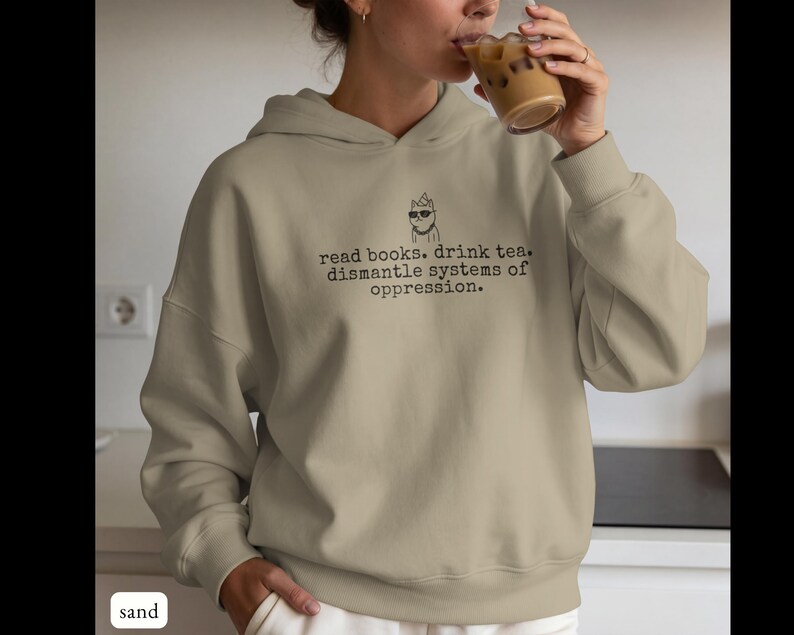 May include: A sand-colored hooded sweatshirt featuring a black graphic of a cat wearing sunglasses and a party hat, accompanied by the text: read books. drink tea. dismantle systems of oppression. The hoodie has long sleeves and ribbed cuffs.