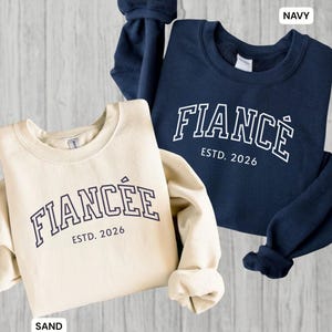 May include: Two sweatshirts, one sand-colored and one navy blue, are displayed. The sand-colored sweatshirt has the word "FIANC&Eacute;E" and "ESTD. 2026" printed on it. The navy blue sweatshirt has the word "FIANC&Eacute;" and "ESTD. 2026" printed on it.