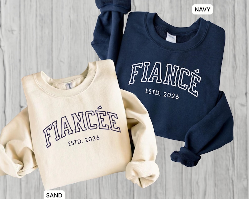 May include: Two sweatshirts, one sand-colored and one navy blue, are displayed. The sand-colored sweatshirt has the word "FIANC&Eacute;E" and "ESTD. 2026" printed on it. The navy blue sweatshirt has the word "FIANC&Eacute;" and "ESTD. 2026" printed on it.