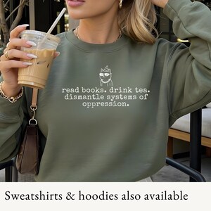 May include: An olive green crewneck sweatshirt featuring a white line-art graphic of a cat wearing a party hat, sunglasses, and a chain, with the text: read books. drink tea. dismantle systems of oppression. The sweatshirt has long sleeves.