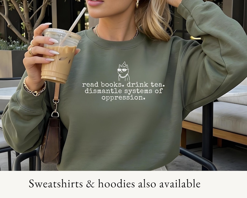 May include: An olive green crewneck sweatshirt featuring a white line-art graphic of a cat wearing a party hat, sunglasses, and a chain, with the text: read books. drink tea. dismantle systems of oppression. The sweatshirt has long sleeves.