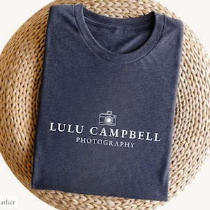 May include: Dark heather gray t-shirt with the text "LULU CAMPBELL PHOTOGRAPHY" and a camera graphic in white. The shirt is folded on a woven, light brown surface.