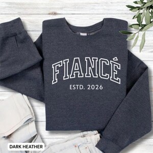 May include: Dark heather gray sweatshirt with the word "FIANC&Eacute;" in white, arched lettering, and "ESTD. 2026" below. The sweatshirt is folded, with the sleeves and bottom hem visible. The words "DARK HEATHER" are at the bottom.