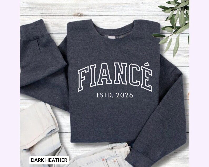 May include: Dark heather gray sweatshirt with the word "FIANC&Eacute;" in white, arched lettering, and "ESTD. 2026" below. The sweatshirt is folded, with the sleeves and bottom hem visible. The words "DARK HEATHER" are at the bottom.