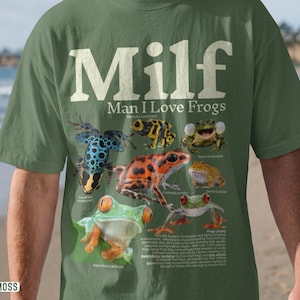 May include: Moss green t-shirt with the text "MILF Man I Love Frogs" in white. The shirt features colorful illustrations of various frog species, including a blue and black frog, and a red and orange frog.