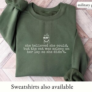 May include: A military green crewneck sweatshirt featuring a white embroidered graphic of a cat wearing sunglasses and a party hat, with the text: she believed she could, but the cat was asleep on her lap so she didn't. The color is military green.