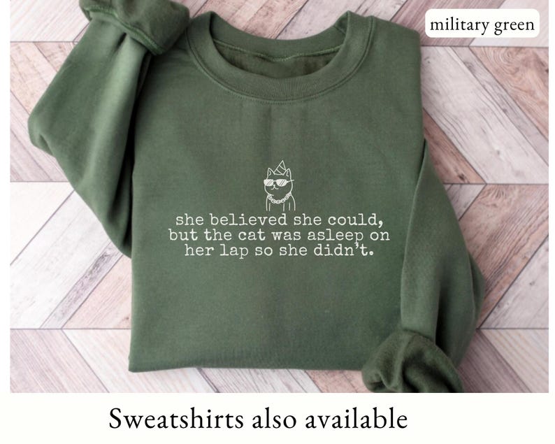 May include: A military green crewneck sweatshirt featuring a white embroidered graphic of a cat wearing sunglasses and a party hat, with the text: she believed she could, but the cat was asleep on her lap so she didn't. The color is military green.