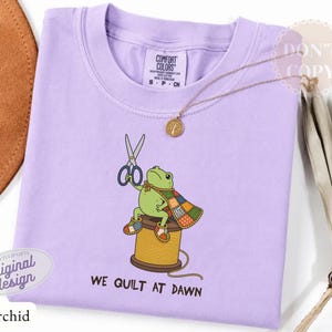 May include: A light purple t-shirt featuring a graphic of a green frog wearing a patchwork cape, sitting on a yellow spool of thread while holding scissors. The text below the frog reads, "WE QUILT AT DAWN." The shirt has a Comfort Colors label.