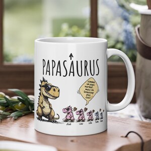 Puede incluir: Taza de cer&aacute;mica blanca con el texto "PAPASAURUS" y un dinosaurio de dibujos animados con gafas de sol. La taza presenta una familia de dinosaurios en rosa y el texto "comme un papa normal mais en beaucoup plus cool!"