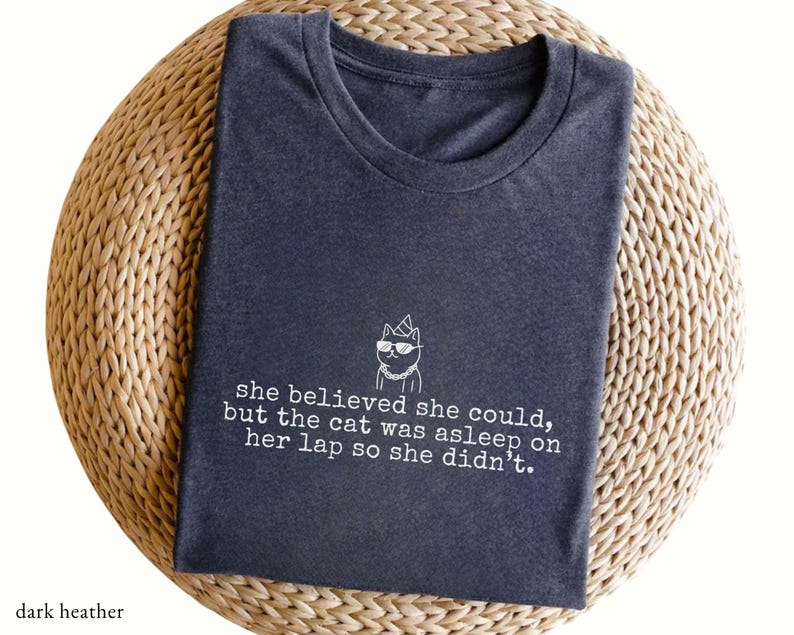 May include: A dark heather gray crew neck t-shirt featuring a white line art graphic of a cat wearing sunglasses and a party hat. Below the cat, the text reads: she believed she could, but the cat was asleep on her lap so she didn't.