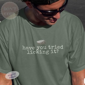 Könnte beinhalten: Ein moosgrünes Kurzarm-T-Shirt mit einer kleinen Grafik eines grauen Steins über dem weißen Text, der lautet, have you tried licking it? Das T-Shirt hat einen Rundhalsausschnitt und eine Standardpassform mit einem schlichten Design.