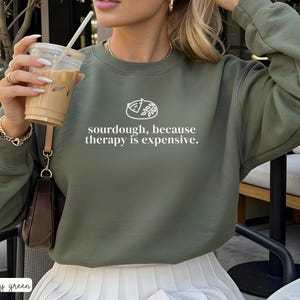 Może przedstawiać: Zielona wojskowa bluza z napisem "sourdough, because therapy is expensive". Bluza ma białą grafikę bochenka chleba. Modelka trzyma przezroczysty plastikowy kubek ze słomką i mrożoną kawą.