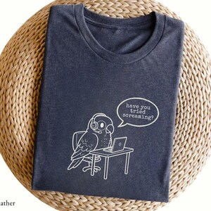 May include: A dark heather gray t-shirt featuring a white line art graphic of a parrot wearing a headset while sitting at a desk with a laptop. A speech bubble above the parrot contains the text, have you tried screaming?