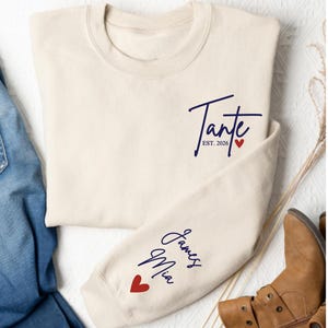 May include: Cream-colored sweatshirt with the word "Tante" in navy blue script, "Est. 2026", and a red heart. The sleeve features the names "James Mia" in navy blue script and a red heart. The sweatshirt is folded.