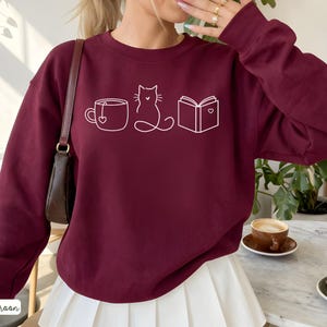 May include: Maroon sweatshirt with a white line drawing of a teacup, cat, and book. The sweatshirt has long sleeves and a crew neck. The word "maroon" is printed in white at the bottom left corner.
