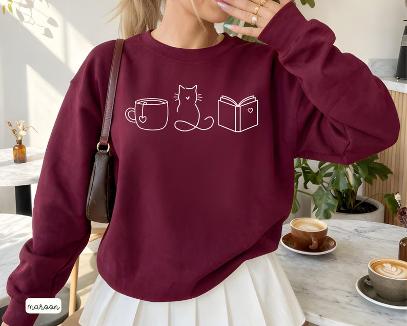 May include: Maroon sweatshirt with a white line drawing of a teacup, cat, and book. The sweatshirt has long sleeves and a crew neck. The word "maroon" is printed in white at the bottom left corner.