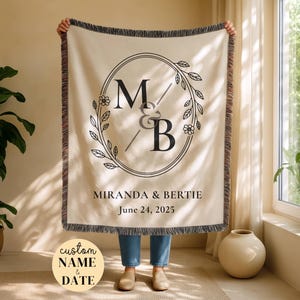 May include: Cream-colored woven throw blanket with a black floral wreath design and the initials "M&B". The blanket also features the names "Miranda & Bertie" and the date "June 24, 2025". The blanket has a fringed edge.