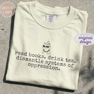 May include: A cream-colored short-sleeve t-shirt featuring a black line drawing of a cat wearing a party hat, sunglasses, and a chain. Below the cat, the text reads: read books. drink tea. dismantle systems of oppression. The shirt has a crew neck.