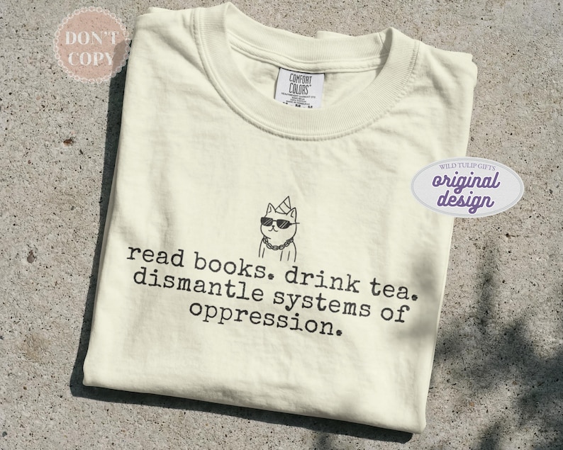 May include: A cream-colored short-sleeve t-shirt featuring a black line drawing of a cat wearing a party hat, sunglasses, and a chain. Below the cat, the text reads: read books. drink tea. dismantle systems of oppression. The shirt has a crew neck.