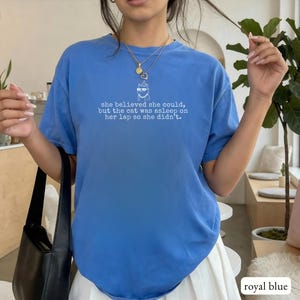 May include: A royal blue short-sleeve t-shirt featuring a white graphic of a cat wearing glasses and the text: "she believed she could, but the cat was asleep on her lap so she didn't." The shirt has a crew neckline and a relaxed fit.