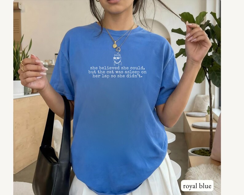May include: A royal blue short-sleeve t-shirt featuring a white graphic of a cat wearing glasses and the text: "she believed she could, but the cat was asleep on her lap so she didn't." The shirt has a crew neckline and a relaxed fit.