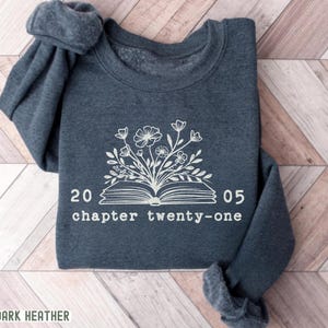 May include: Dark heather gray sweatshirt with a white floral and book design. The design includes the text "20 05 chapter twenty-one". The words "DARK HEATHER" are printed at the bottom of the sweatshirt.