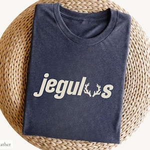May include: Dark heather blue t-shirt with the word "jegulus" in white, featuring antler-like designs. The shirt is folded and placed on a woven, circular surface.