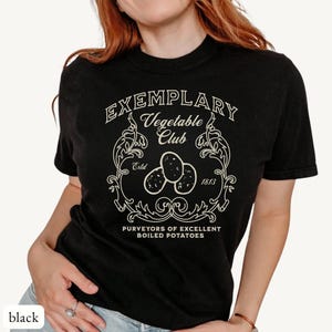 Op de afbeelding: Zwart t-shirt met een crèmekleurige print. De print luidt "Exemplary Vegetable Club Estd 1813" met een illustratie van drie aardappelen. Daaronder staat "Purveyors of Excellent Boiled Potatoes."