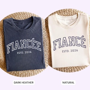 May include: Two t-shirts, one dark heather and one natural, each with the word "FIANC&Eacute;E" arched across the front in white block letters, with "ESTD. 2026" below. The shirts are folded on a woven mat.