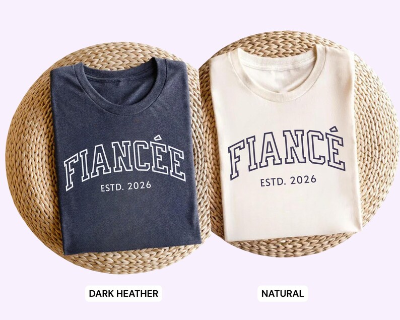 May include: Two t-shirts, one dark heather and one natural, each with the word "FIANC&Eacute;E" arched across the front in white block letters, with "ESTD. 2026" below. The shirts are folded on a woven mat.