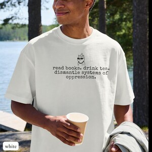 May include: A white short-sleeved t-shirt featuring a black graphic of a cat wearing sunglasses and a party hat, with the text: read books. drink tea. dismantle systems of oppression. The shirt has a crew neckline and a relaxed fit.