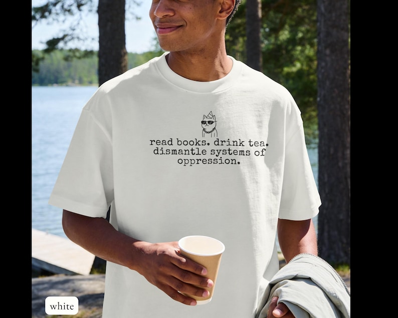 May include: A white short-sleeved t-shirt featuring a black graphic of a cat wearing sunglasses and a party hat, with the text: read books. drink tea. dismantle systems of oppression. The shirt has a crew neckline and a relaxed fit.