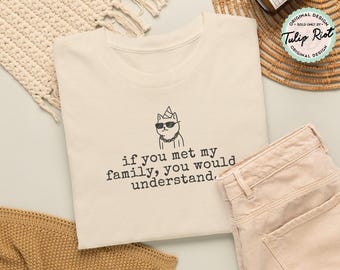 Può includere: Una t-shirt color crema con un disegno a linee nere di un gatto che indossa occhiali da sole, un cappellino da festa e una collana a catena. Sotto il gatto, il testo recita: if you met my family, you would understand. La maglietta è piegata.