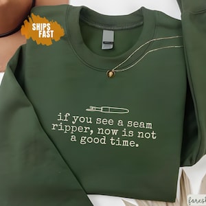 Könnte beinhalten: Forstgrünes Sweatshirt mit dem Text "if you see a seam ripper, now is not a good time." Eine goldene Halskette mit Anhänger liegt auf dem Sweatshirt. Ein brauner Hut ist in der oberen linken Ecke sichtbar.