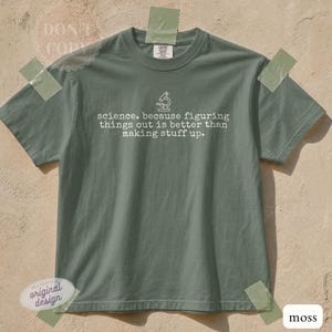 Puede incluir: Una camiseta de manga corta color verde musgo con un icono de línea blanca de un microscopio sobre el texto: science. because figuring things out is better than making stuff up. La camiseta tiene cuello redondo y un diseño de corte estándar.