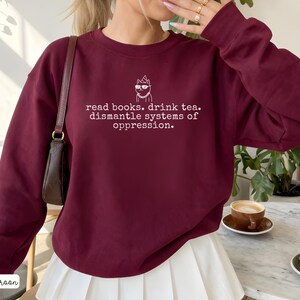 May include: A maroon long-sleeve crewneck sweatshirt featuring a white graphic of a cat wearing sunglasses and a party hat, with the text: read books. drink tea. dismantle systems of oppression. The sweatshirt has a classic fit and ribbed cuffs.