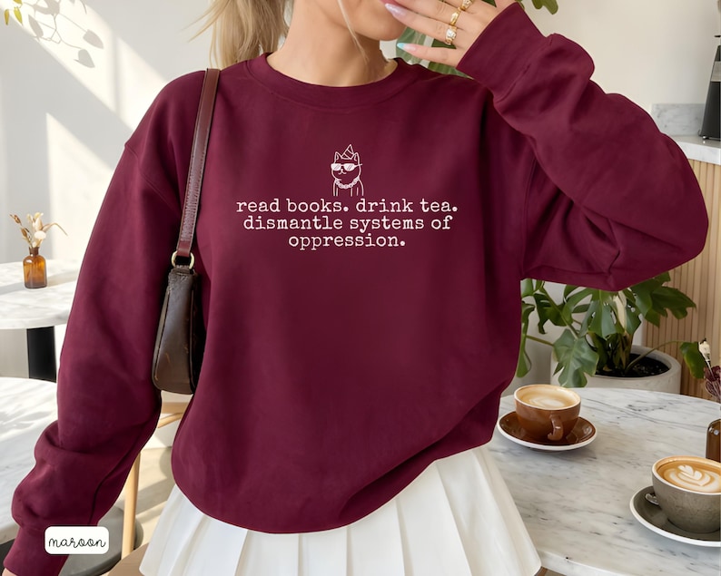 May include: A maroon long-sleeve crewneck sweatshirt featuring a white graphic of a cat wearing sunglasses and a party hat, with the text: read books. drink tea. dismantle systems of oppression. The sweatshirt has a classic fit and ribbed cuffs.