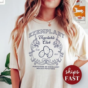 Può includere: T-shirt color crema con grafica blu navy. La grafica recita "Exemplary Vegetable Club" con un'illustrazione di patate e decorazioni. È incluso anche il testo "Purveyors of Excellent Boiled Potatoes".