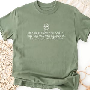 May include: An olive green short-sleeved t-shirt featuring a white line-art graphic of a cat wearing sunglasses and a party hat, with the text: she believed she could, but the cat was asleep on her lap so she didn't.