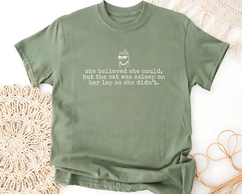 May include: An olive green short-sleeved t-shirt featuring a white line-art graphic of a cat wearing sunglasses and a party hat, with the text: she believed she could, but the cat was asleep on her lap so she didn't.