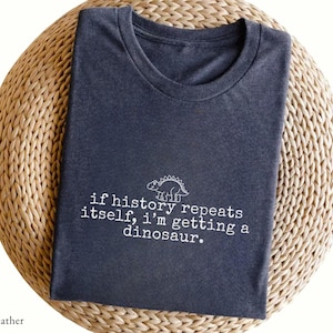 Peut inclure: Un t-shirt à col rond gris chiné foncé avec une illustration au trait blanc d'un dinosaure stégosaure au-dessus du texte, "if history repeats itself, i'm getting a dinosaur." Le t-shirt est plié soigneusement pour montrer le motif.