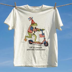 Könnte beinhalten: Weißes T-Shirt mit einem Cartoon-Frosch auf einem Roller, der mit der amerikanischen Flagge verziert ist. Der Frosch trägt einen patriotischen Hut und eine Jacke. Der Text "Off to commit patriotic tomfoolery" ist darunter gedruckt.