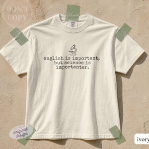 May include: An ivory short-sleeve t-shirt featuring a black line-art illustration of a microscope centered above the text, "english is important, but science is importanter." The shirt has a crew neckline and a standard fit, presented in a neutral tone.